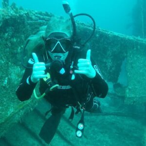 A female scuba diver in black gear giving two thumbs up underwater in front of a heavily encrusted shipwreck structure.