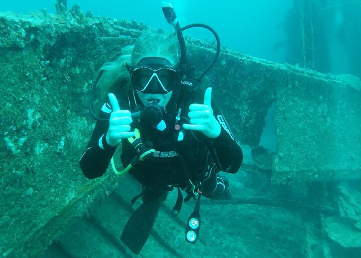 A female scuba diver in black gear giving two thumbs up underwater in front of a heavily encrusted shipwreck structure.