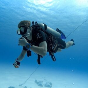 A scuba diver floating in the clear blue water, gripping a rope descending to the sandy bottom, with bright sunlight filtering from above.
