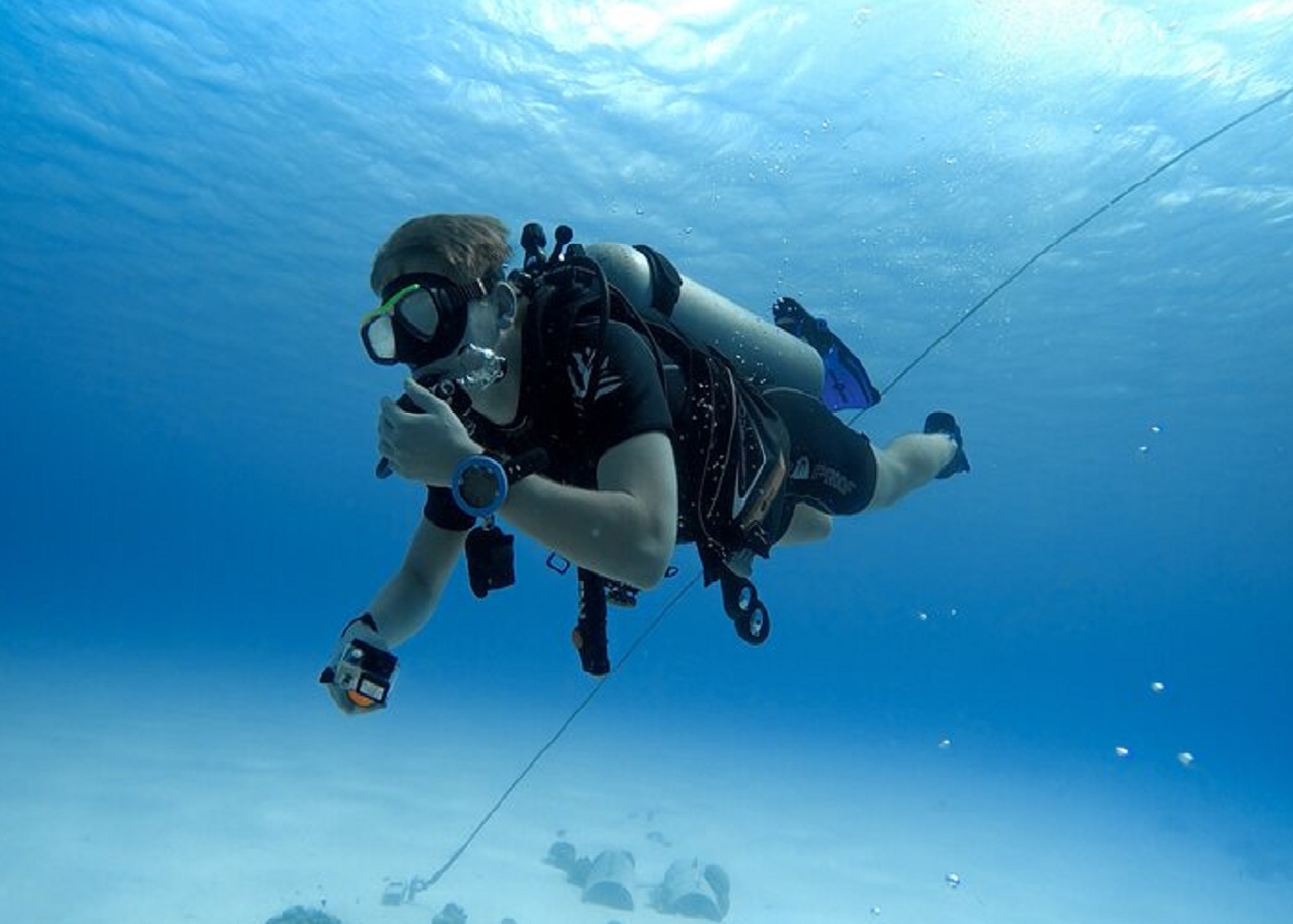 A scuba diver floating in the clear blue water, gripping a rope descending to the sandy bottom, with bright sunlight filtering from above.