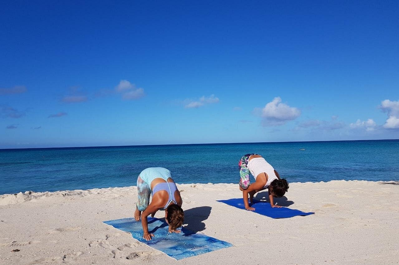 Two women practicing Beach Yoga in Crow Pose on blue mats on a white sand beach with turquoise ocean and blue sky in the background, demonstrating balance and strength.