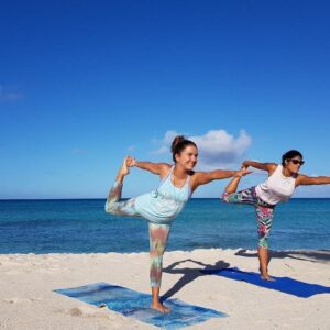 Two women demonstrating a balanced Dancer's Pose (Natarajasana) on blue mats during daytime Beach Yoga with a clear blue sky and turquoise ocean.