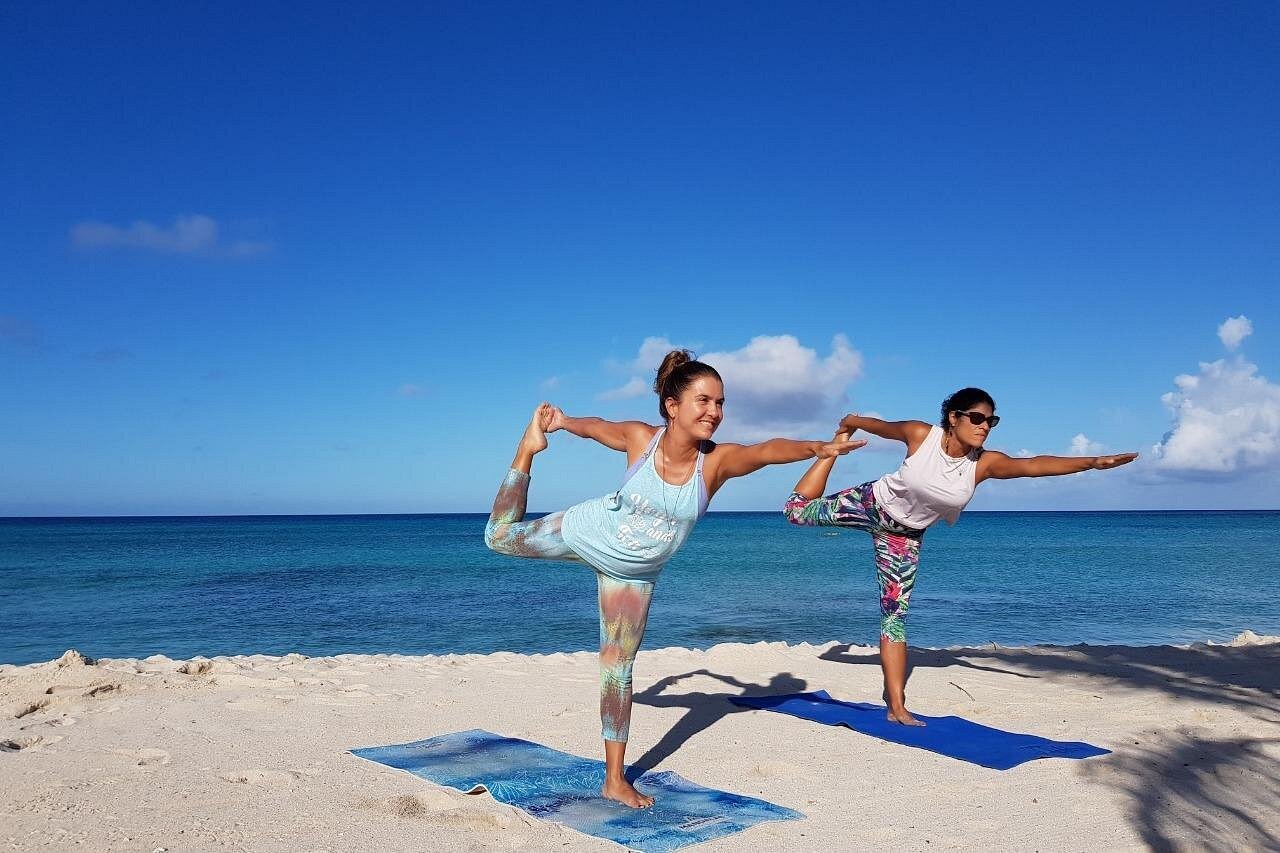 Two women demonstrating a balanced Dancer's Pose (Natarajasana) on blue mats during daytime Beach Yoga with a clear blue sky and turquoise ocean.
