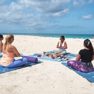 A group of four women seated in a circle on blue yoga mats on the white sand of Eagle beach, participating in a guided Meditation and Chakras opening session. The main instructor sits facing the group.