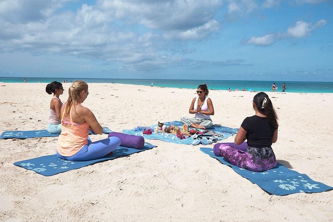 A group of four women seated in a circle on blue yoga mats on the white sand of Eagle beach, participating in a guided Meditation and Chakras opening session. The main instructor sits facing the group.