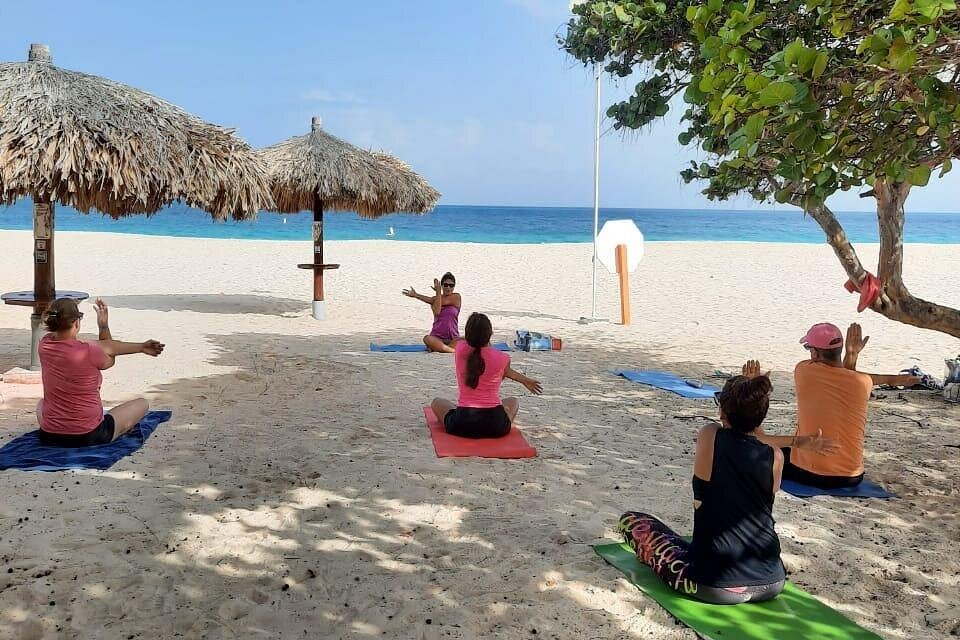 A group seated on yoga mats, gently stretching their arms under the shade of a tree, preparing for meditation or sound bath on the beach. All levels welcome to this calming setting.
