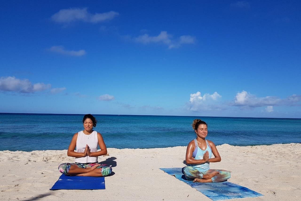 Two women sitting in Easy Pose (Sukhasana) on yoga mats, eyes closed, palms together, engaged in deep meditation or sound bath on the tranquil beach.