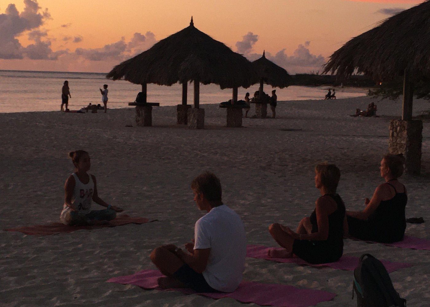 A yoga instructor leading a small group in a seated pose on the sand at sunset, creating a serene environment for meditation or Sound Bath. All levels welcome.