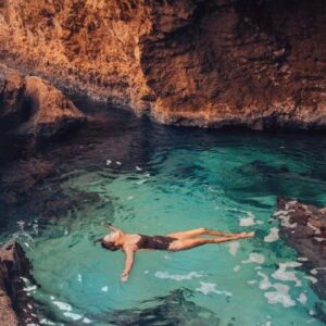 A person floating on their back in the clear, turquoise water of a secluded Natural Pool surrounded by towering, warm-toned rock formations, highlighting the chance to Swim in Natural Pool and Escape the Heat.