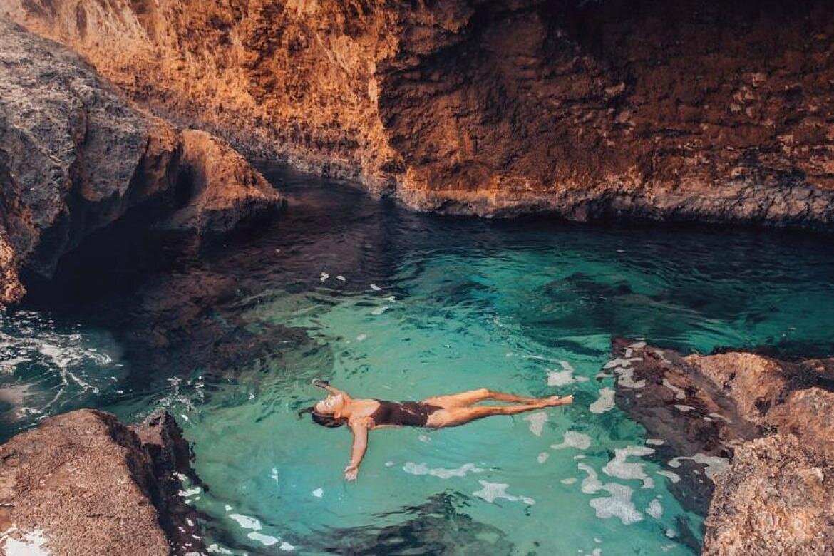 A person floating on their back in the clear, turquoise water of a secluded Natural Pool surrounded by towering, warm-toned rock formations, highlighting the chance to Swim in Natural Pool and Escape the Heat.