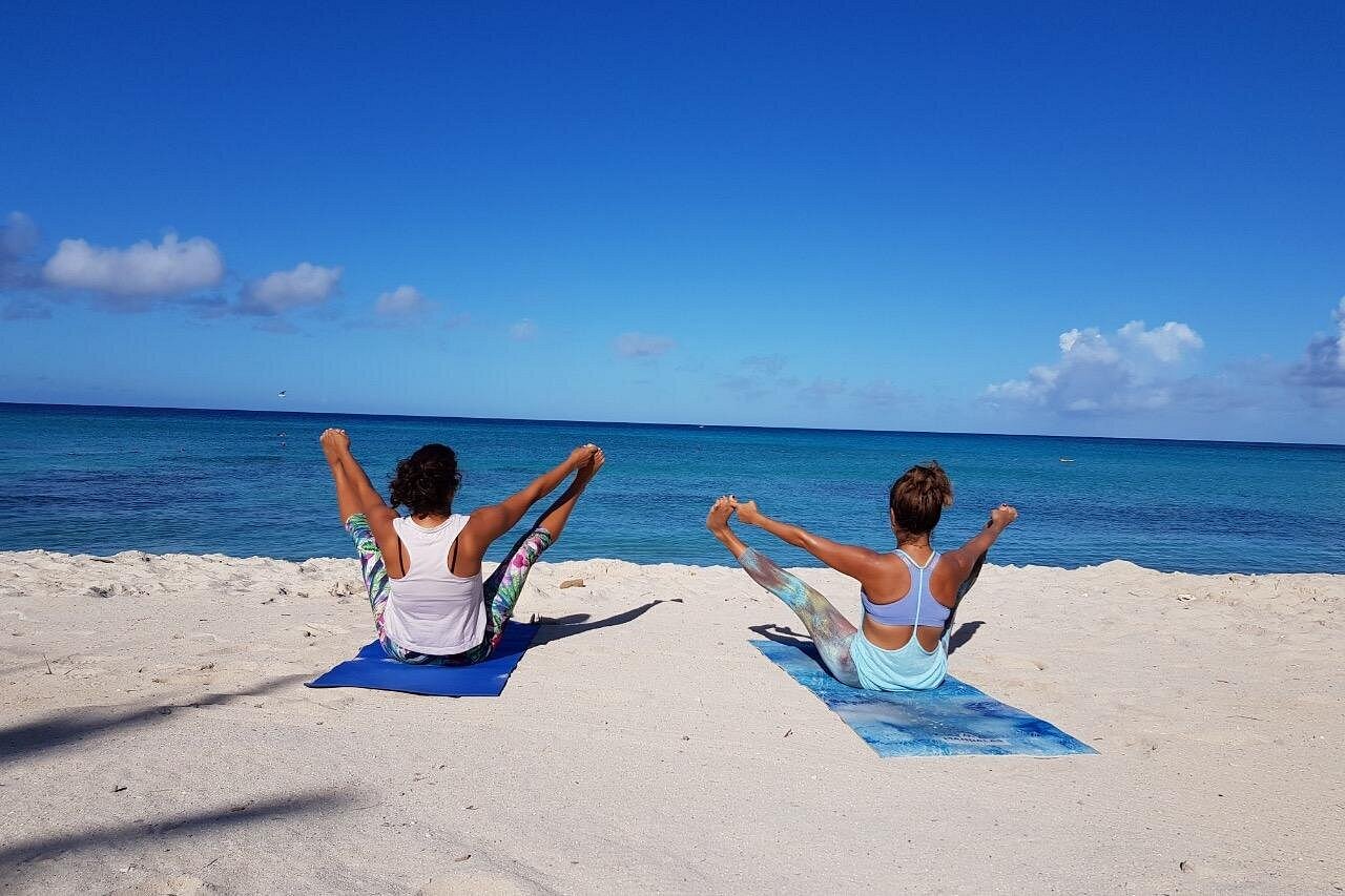 Two women seated on yoga mats facing the ocean, practicing a variation of Upavistha Konasana (Seated Wide-Legged Forward Bend), excellent for all levels welcome to a relaxing meditation session.