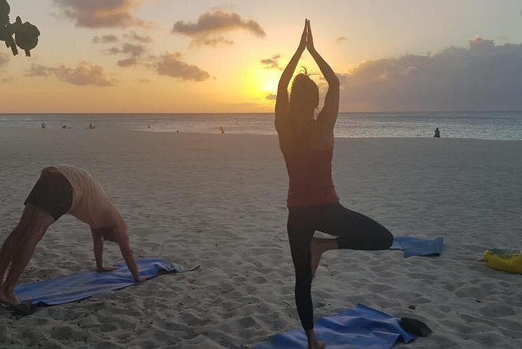 A woman in Tree Pose (Vrksasana) and a man in Downward-Facing Dog (Adho Mukha Svanasana) practicing Beach Yoga against a gorgeous sunset backdrop, perfect for pre-meditation flow.