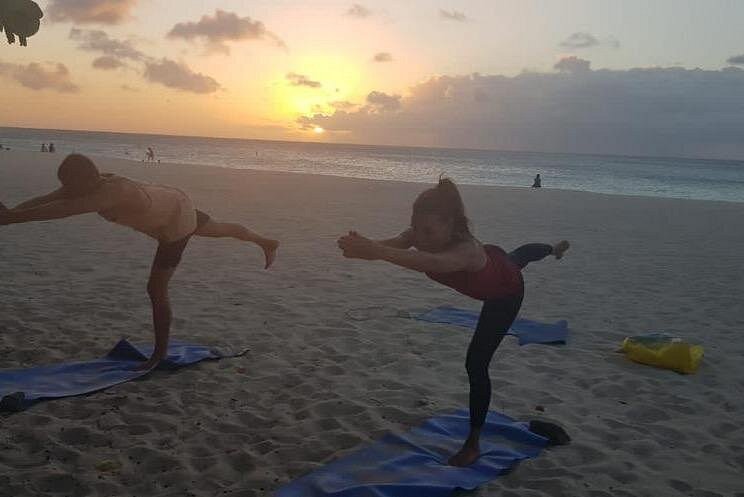 Two people holding the balanced Warrior III Pose during a tranquil sunset Beach Yoga session, promoting focus before a Sound Bath & meditation.