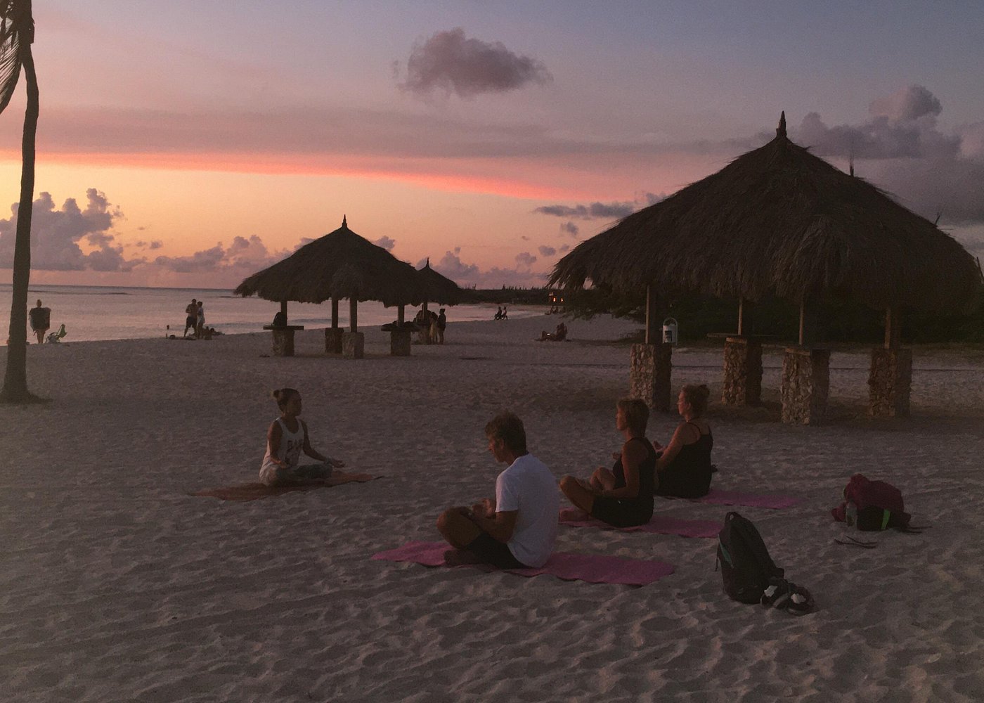A small group sitting cross-legged in silent meditation on pink yoga mats on the beach at sunset, with palapas providing shade and a calm atmosphere for a Sound Bath.