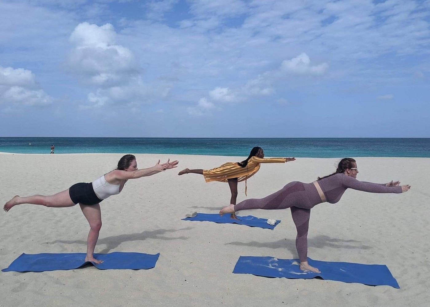 Three people practicing the challenging Warrior III pose during Beach Yoga, emphasizing focus, a key element of meditation and sound bath practice, on a sunny day by the ocean.