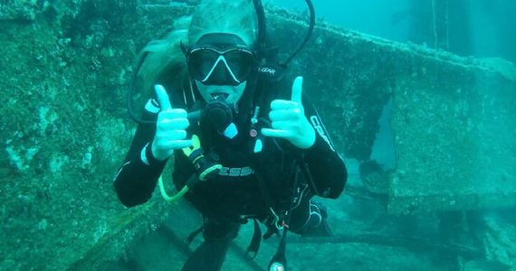 A female scuba diver in black gear giving two thumbs up underwater in front of a heavily encrusted shipwreck structure.