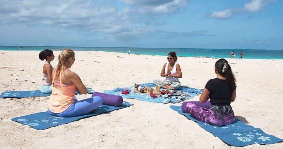 A group of four women seated in a circle on blue yoga mats on the white sand of Eagle beach, participating in a guided Meditation and Chakras opening session. The main instructor sits facing the group.