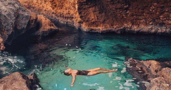 A person floating on their back in the clear, turquoise water of a secluded Natural Pool surrounded by towering, warm-toned rock formations, highlighting the chance to Swim in Natural Pool and Escape the Heat.