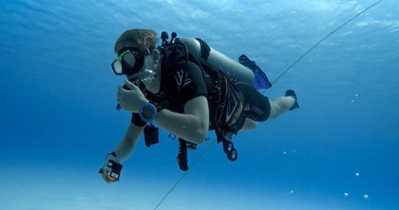 A scuba diver floating in the clear blue water, gripping a rope descending to the sandy bottom, with bright sunlight filtering from above.